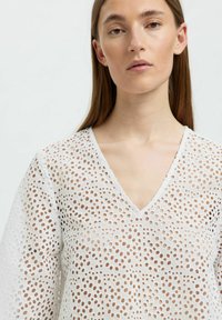 Hvid bluse med V-udskæring, der har et tekstureret design af cirkulære udskæringer, lange ærmer og et let stof.