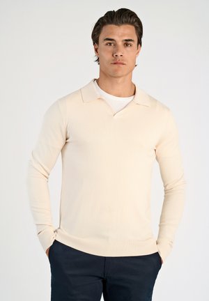Lichtbeige poloshirt met lange mouwen en V-hals, met ribgebreide mouwen, gedragen over een witte T-shirt, gecombineerd met donkerblauwe broeken.
