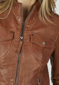 Giacca di pelle marrone con un design attillato, dotata di una zip frontale, due tasche sul petto e dettagli cuciti sulle spalle.