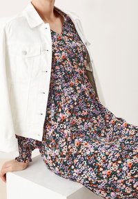 Valkoinen farkkutakki kerroksittain mustan kukkamekon päällä, jossa on purppuran, oranssin ja vihreän värisiä kuvioita. Mekossa on röyhelökoristeet hihoissa ja vyötäröllä.