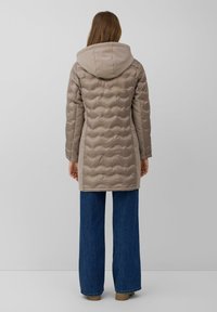 Chaqueta acolchada beige con capucha, con una textura de acolchado ondulado; combinada con unos vaqueros anchos azules y calzado marrón claro.