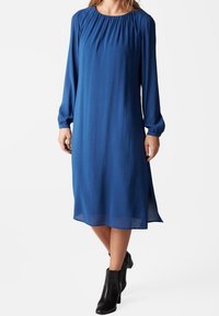 Abito blu a maniche lunghe con scollatura arricciata, tessuto fluido, lunghezza al ginocchio e spacchi laterali, abbinato a stivali neri alla caviglia.