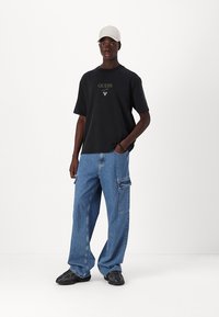 Svart bomull T-skjorte med sentralt logo, kombinert med blå denim cargo-bukser med lommer. Brukt med svarte joggesko og en lys caps.