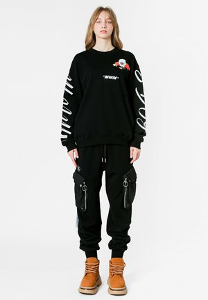Sudadera negra con diseño gráfico y texto blanco, combinada con pantalones cargo negros con bolsillos con cremallera. Botas color beige completan el conjunto.