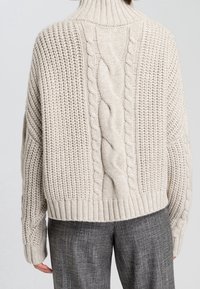 Kermaisen värinen paksu neulepaita, jossa on korkea kaulus, punottu keskiosa ja resorireunus, yhdistetty harmaisiin ruudullisiin housuihin.