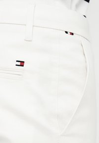Pantalon blanc avec passants de ceinture, poches avant et arrière, avec une petite broderie de logo rectangulaire rouge, blanc et noir.