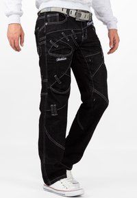 Jeans in denim nero con cuciture bianche, multiple tasche e dettagli di taglio unici. Abbinati a una cintura grigia e sneakers bianche.