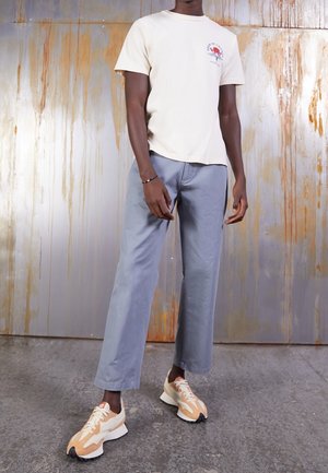 Henkilö, jolla on valkoinen kuvioitu t-paita, vaaleansiniset leveälahkeiset housut ja beige-valkoiset lenkkitossut, ruosteen värjäämää metallitaustaa vasten.