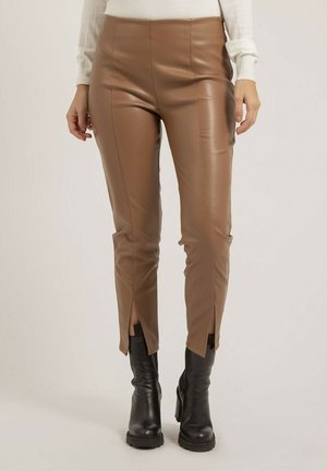 Femme portant un pantalon en cuir marron avec des fentes à l'avant des jambes et des bottines noires, debout devant un fond clair uni.