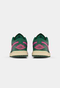 Ténis verdes em pele com uma faixa de destaque rosa e logotipo bordado na parte de trás, acompanhados de uma sola de borracha bege.