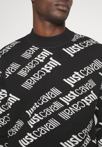 Närbild av en svart tröja med vit text "Just Cavalli" som upprepas diagonalt över tyget, buren av en person med mörk hud.