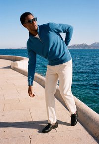 Mann mit Sonnenbrille, der einen blauen Pullover und weiße Hose trägt, lehnt sich zurück auf einer sonnigen Meerespromenade mit Bergen im Hintergrund.