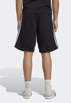 Persona de espaldas vestida con pantalones cortos negros con franjas blancas laterales, calcetines blancos a rayas y sandalias blancas tipo slide sobre un fondo liso.