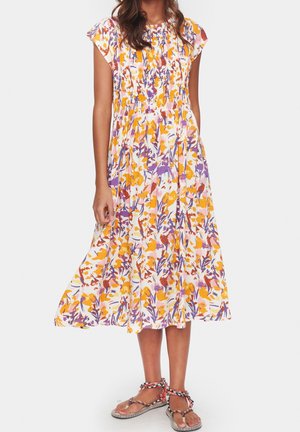 Kobieta nosząca luźną sukienkę o długości midi z krótkimi rękawami i kolorowym, kwiatowym wzorem w żółtym, fioletowym i pomarańczowym kolorze, w połączeniu z plecionymi sandałami.