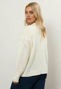 En blond kvinna iförd en vit stickad tröja med flätade mönster på ärmarna, som tittar över sin axel mot en enkel ljus bakgrund.