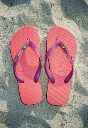 Paire de tongs Havaianas roses et violettes avec logo du drapeau brésilien reposant sur une plage de sable.