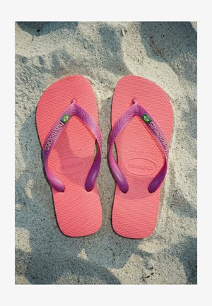 Paire de tongs Havaianas roses et violettes avec logo du drapeau brésilien reposant sur une plage de sable.