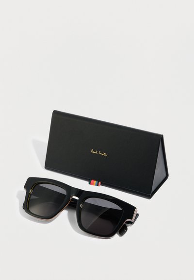 Μαύρα υπερμεγέθη γυαλιά ηλίου με σκούρους φακούς, με πολύχρωμο ριγωτό διακοσμητικό στο μπράτσο, παρουσιάζονται μέσα σε μαύρη τριγωνική θήκη.