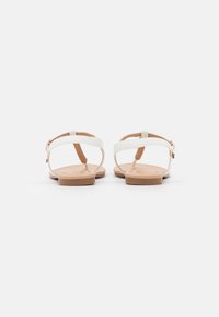 Infradito bianchi piatti con design a T, suola beige e fibbia regolabile alla caviglia. Texture liscia con uno stile minimalista e senza motivi visibili.