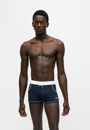Homme athlétique debout, face à l’objectif, portant un short de bain bleu marine avec une ceinture blanche et un passepoil, sur un fond clair uni.
