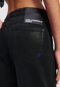 Crne traperice od denima s vidljivom crnom kožnom etiketom s natpisom "KARL LAGERFELD JEANS." Sadrži plavi izvezeni "K" na stražnjem džepu.