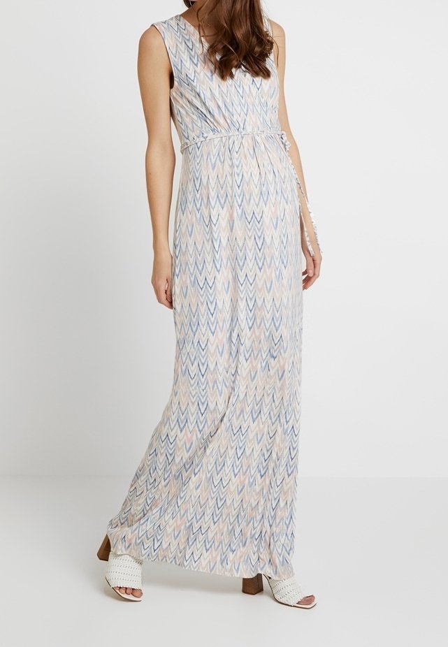 Femme portant une robe maxi sans manches avec un léger motif chevron et des sandales blanches à talons ouverts sur fond uni.