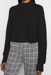 Frau in einem schwarzen, gerippten Strickpullover mit langen Ärmeln und hohem Kragen, kombiniert mit schwarz-weißen karrierten Hosen.