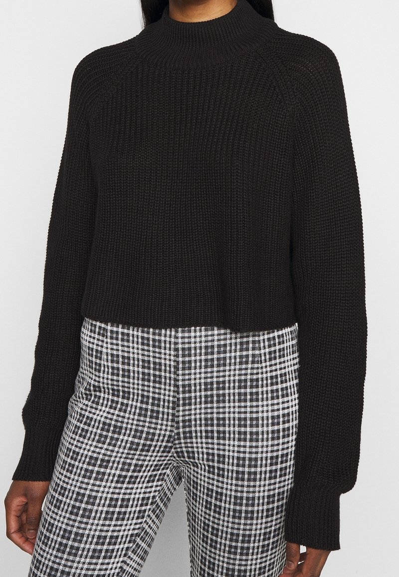 Frau in einem schwarzen, gerippten Strickpullover mit langen Ärmeln und hohem Kragen, kombiniert mit schwarz-weißen karrierten Hosen.