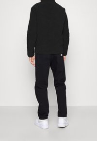 Veste en polaire noire avec un col montant et une finition texturée, associée à un pantalon foncé et des baskets blanches, vue de dos.