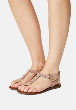 Sandales en suède beige avec un design en T, accentuées de clous dorés et dotées d'une semelle en cuir plate. Ouvertes sur le bout avec une couverture minimale et un style décontracté.