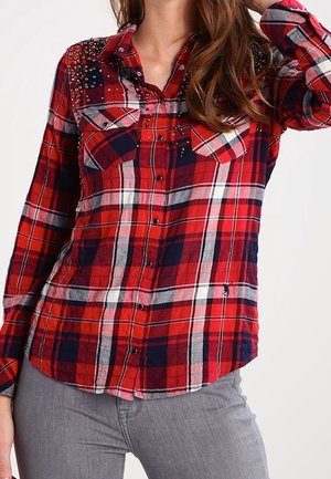 Vrouw draagt een rood, marineblauw en wit geruit overhemd met knopen en studs op de schouders, gecombineerd met een grijze broek.