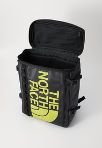 Sac à dos noir en matériau durable, avec un grand logo jaune, un sommet zippé et des sangles latérales ajustables. L'intérieur comprend une poche en filet.