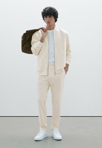 Veste bomber beige clair, pantalon de jogging assorti et baskets blanches. Le modèle tient un sac marron. Tissu lisse, coupe décontractée, design minimaliste.