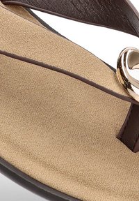 Las chanclas marrones tienen una plantilla texturada en beige, correas marrones y un acento metálico redondo en el tobillo.
