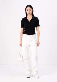 Polo noir à manches courtes, associé à un jean blanc à jambes larges. Le modèle tient un petit sac blanc, se tenant contre un fond uni.
