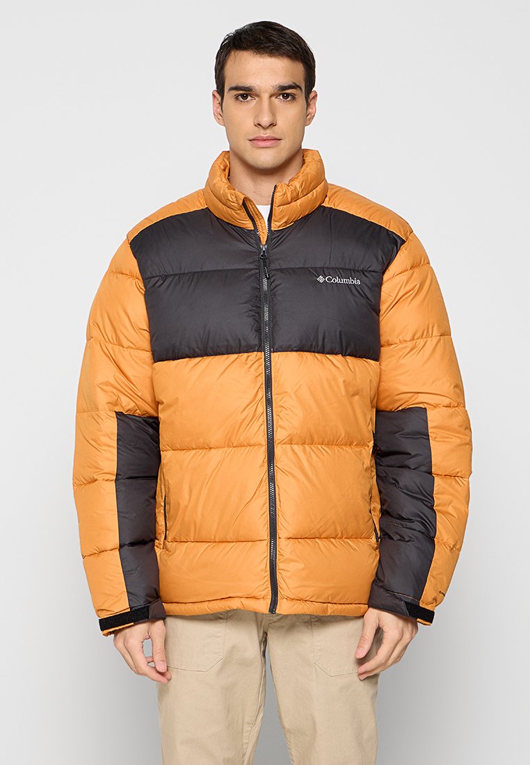 Junger Mann trägt eine orange-schwarze Columbia Daunenjacke mit Reißverschluss und beige Hose, steht vor einem schlichten Hintergrund.