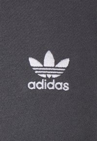 Šedá textilie s texturovaným povrchem, na které je bílá vyšitá značka Adidas skládající se ze tří listů a paralelních pruhů.