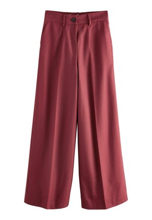 Osoba nosząca wysoki stan ciemnoczerwonych spodni z elastycznym pasem i tylnymi kieszeniami z wypustką, ręka spoczywająca w bocznej kieszeni, widoczny czarny top.