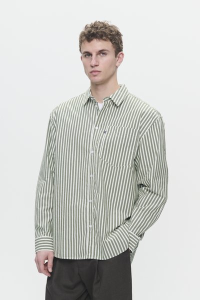 Giovane uomo che indossa una camicia a maniche lunghe a righe verticali verdi e bianche e pantaloni scuri, in piedi con una mano in tasca su uno sfondo semplice.