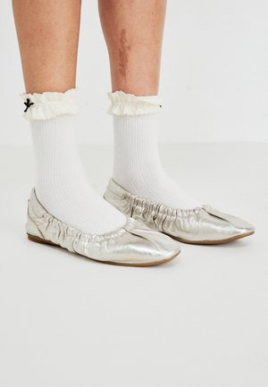 Metāliskas sudraba krāsas baleta kurpes ar salocītām malām, kopā ar baltām ribotām zeķēm ar viļņainu apkakli un melnu loku akcentu.