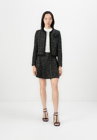 Black and white tweed jacket with matching skirt, textured fabric, and short sleeves. White turtleneck underneath, paired with black strappy heels.