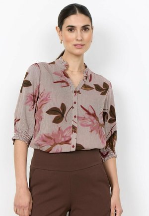 Vrouw met donker haar in een paardenstaart, gekleed in een roze en bruine bloemenblouse met knopen en bruine hoog getailleerde broek, staand tegen een witte achtergrond.