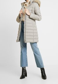 Lichtgrijze pufferjas met een bontgevoerde capuchon, voorzien van een ritsluiting en een doorgestikte textuur. Draag het met lichtblauwe flared jeans en zwarte laarzen.