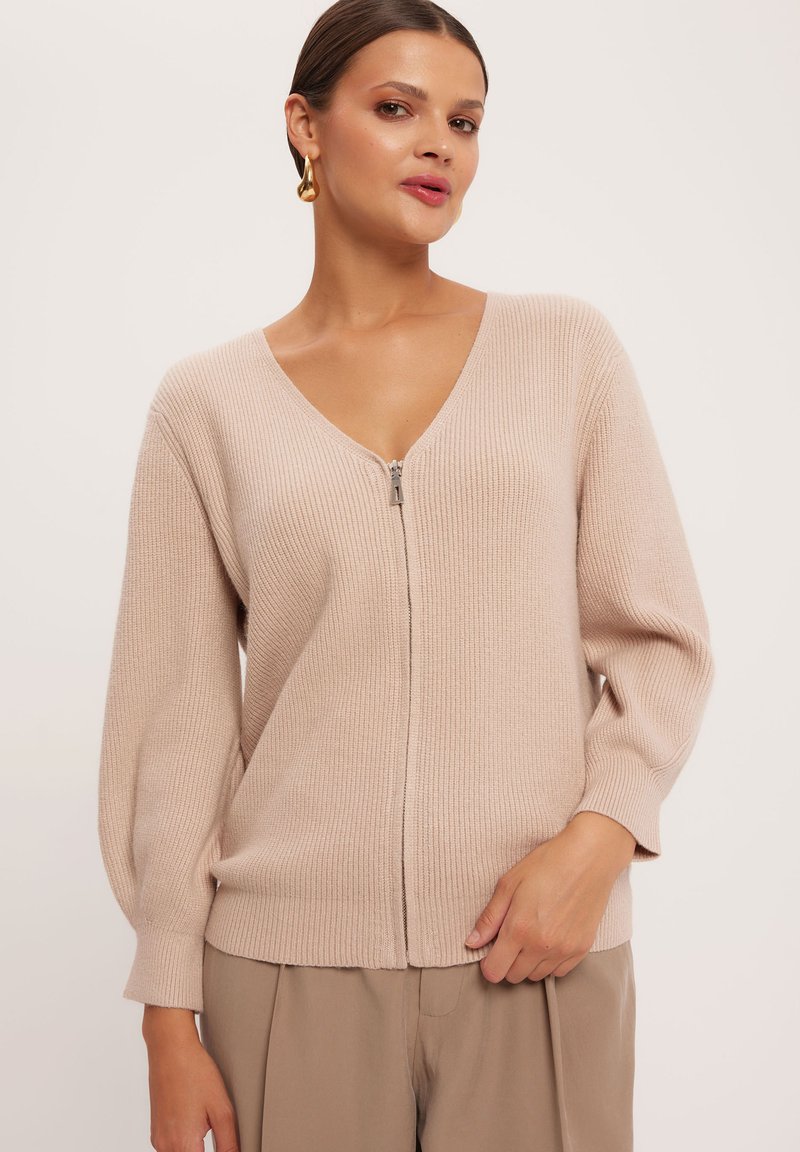 Jasnoróżowy, prążkowany kardigan z dekoltem w kształcie V i zamek błyskawiczny. Posiada długie, lekko bufiaste rękawy oraz gładką, miękką teksturę.