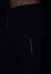 Schwarze sportliche Jogginghosen mit einem elastischen Bund, ausgestattet mit einem reflektierenden Kordelzug und einem vertikalen weißen Logo an der Seite. Glatte Textur.