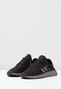 Czarne sportowe buty z siateczkową cholewką o fakturowanej powierzchni, z charakterystyczną podeszwą w kształcie diamentowego wzoru i czarnymi sznurowadłami, zapewniające lekką konstrukcję.