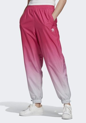 Vrouw draagt Adidas-broek met een overgang van roze naar lichtgrijs, elastische boorden en witte sneakers, handen in de zakken.
