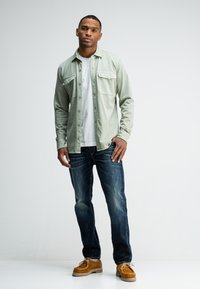 Lichtgroen overhemd met knopen, twee borstzakken, een witte T-shirt eronder, donkere jeans en bruine suède schoenen. Eenvoudige moderne silhouet.