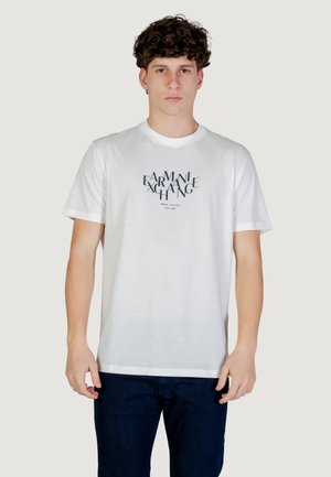 Giovane con capelli ricci che indossa una maglietta bianca Armani Exchange con testo blu navy e jeans blu scuro, in piedi davanti a uno sfondo semplice.