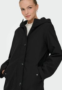 Crna kapućasta jakna od glatke tkanine, s prednjim zatvaračem, četiri dugmeta i bočnim džepovima. Jednostavan, moderan dizajn.